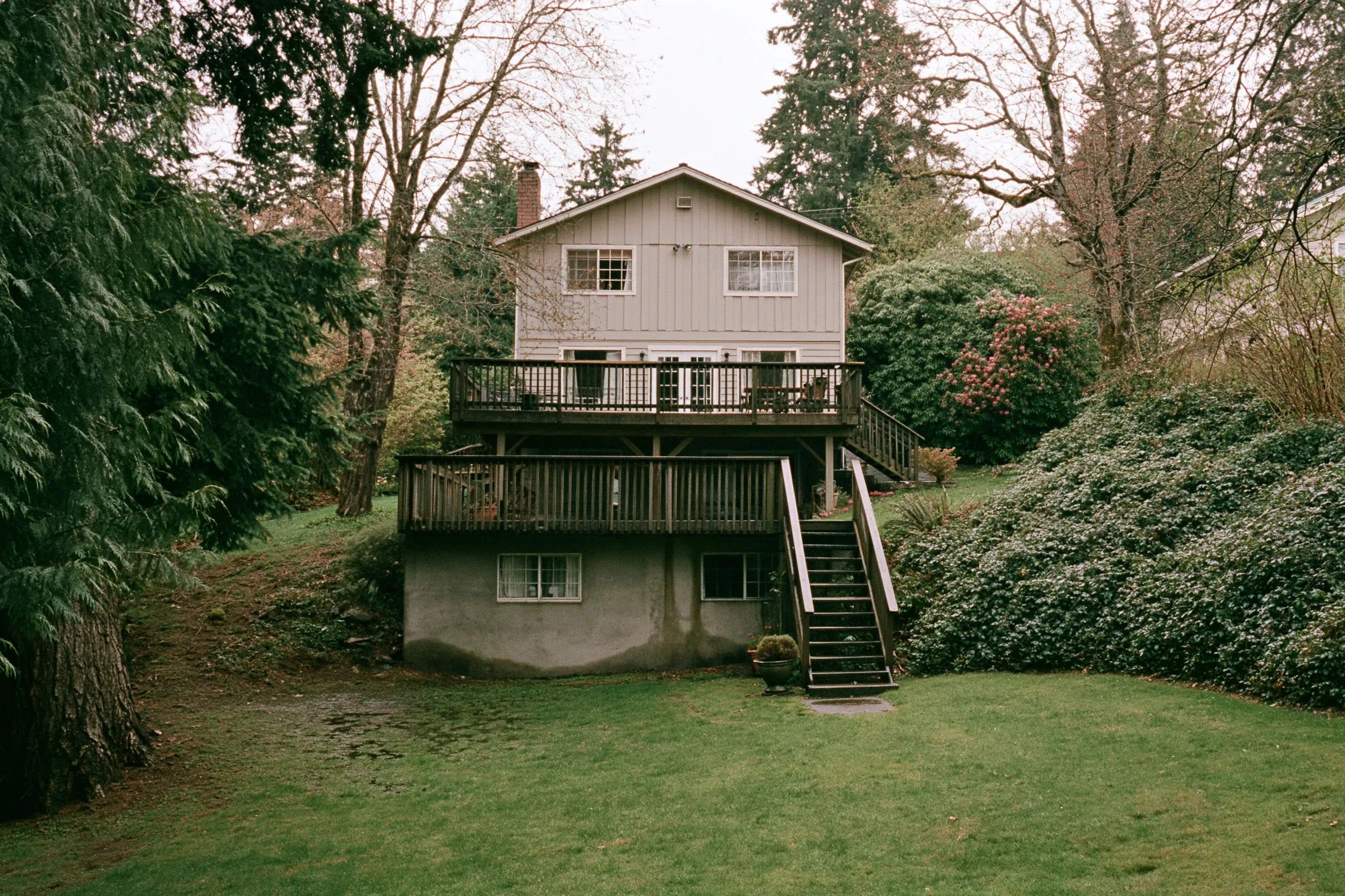 Rear of a two-story house on a gentle slope with a daylight basement, the poured concrete foundation wall showing at the lower level, a deck above and Douglas firs behind a lawn